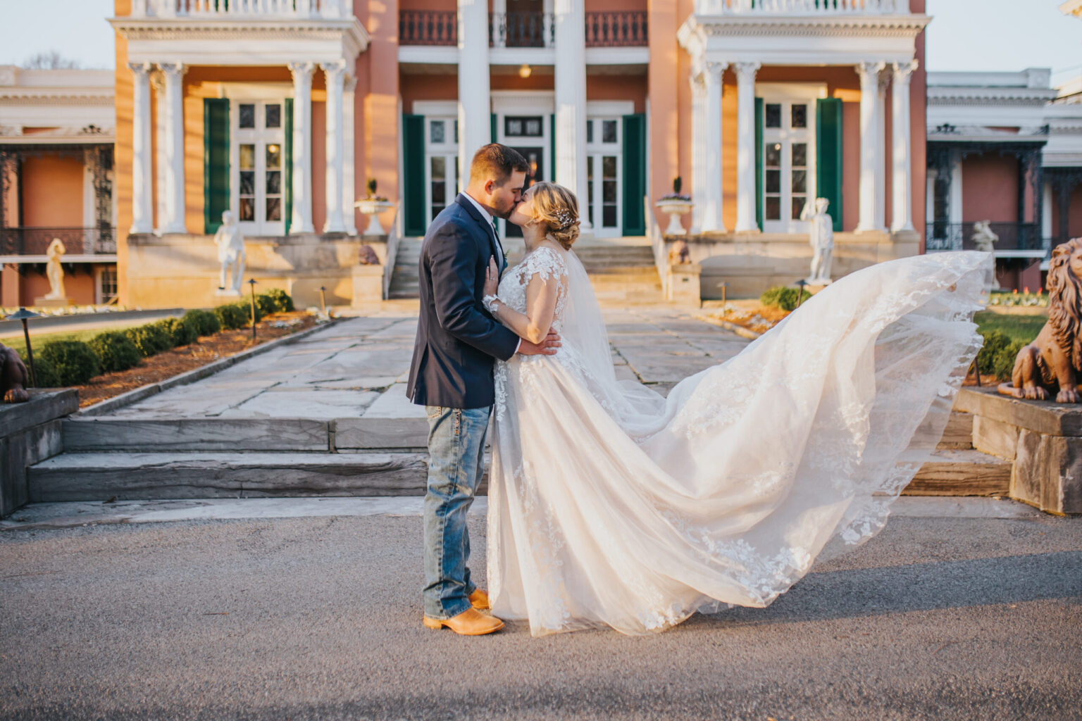 Elopement Wedding A bride and groom share an intimate moment in front of a grand, pink mansion with white columns. The bride, in a flowing white gown, has her back turned slightly, showing her train cascading behind her. The groom is dressed in a dark suit paired with light blue jeans and brown shoes, capturing the essence of an elopement. Elopements Inc