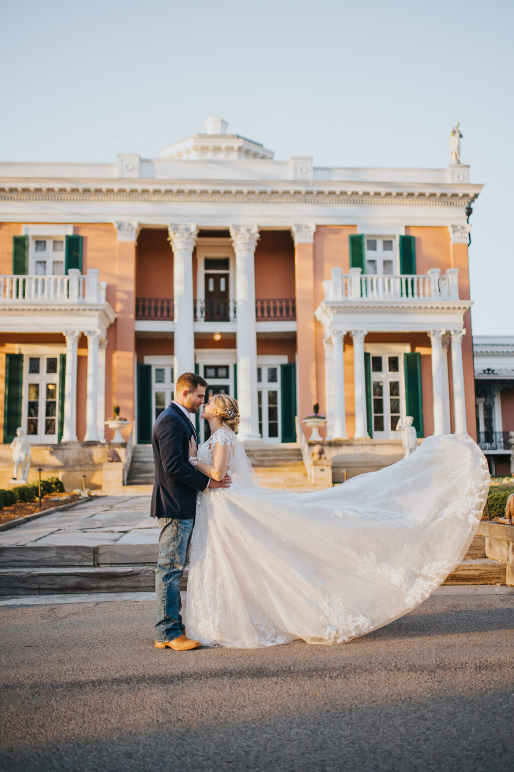 Elopement Wedding A couple stands embracing in front of an elegant, historic building with tall columns and large windows. The bride, eloping in a flowing white gown with lace details, creates a dramatic effect as her dress fans out. The groom complements her perfectly, dressed in a dark suit with brown shoes. Elopements Inc