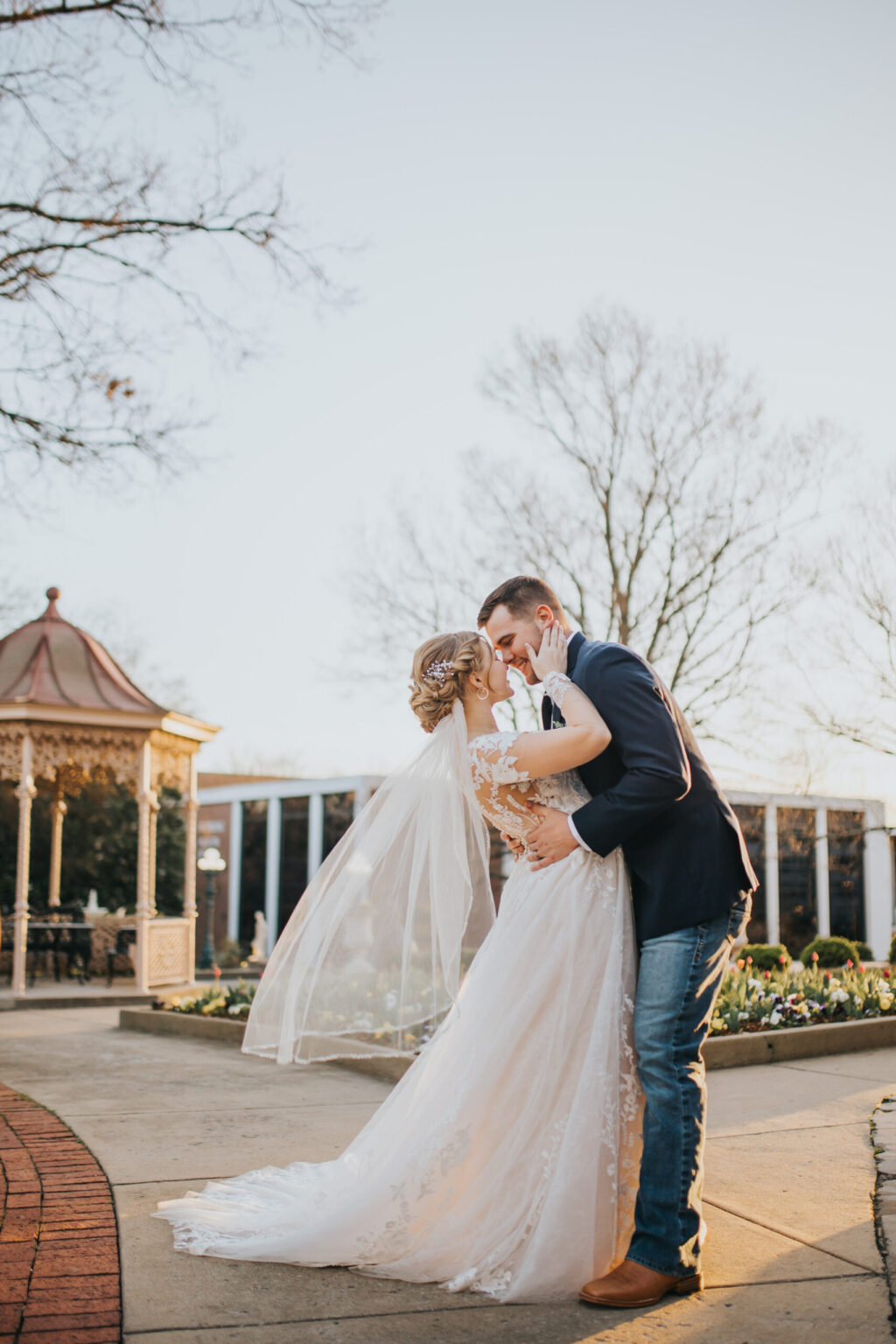 Elopement Wedding A bride and groom share a kiss outdoors in front of a gazebo, celebrating their intimate elopement. The bride wears a long, white dress with a lace top and veil, and the groom is dressed in a navy suit jacket, blue jeans, and brown boots. Trees with bare branches and a clear sky are visible in the background. Elopements Inc