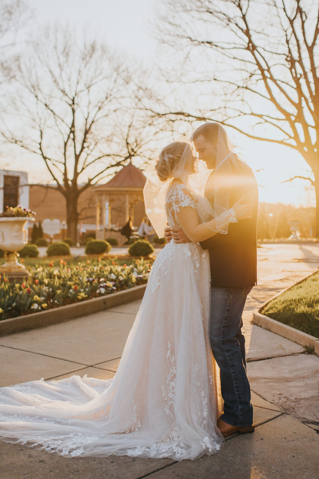 Elopement Wedding A bride in a long lace gown and veil embraces a groom in a dark jacket and jeans. The couple stands on a sunlit pathway surrounded by a garden, with trees and a gazebo in the background. The sun is low, casting a golden glow as they enjoy an intimate moment during their elopement, forehead to forehead. Elopements Inc