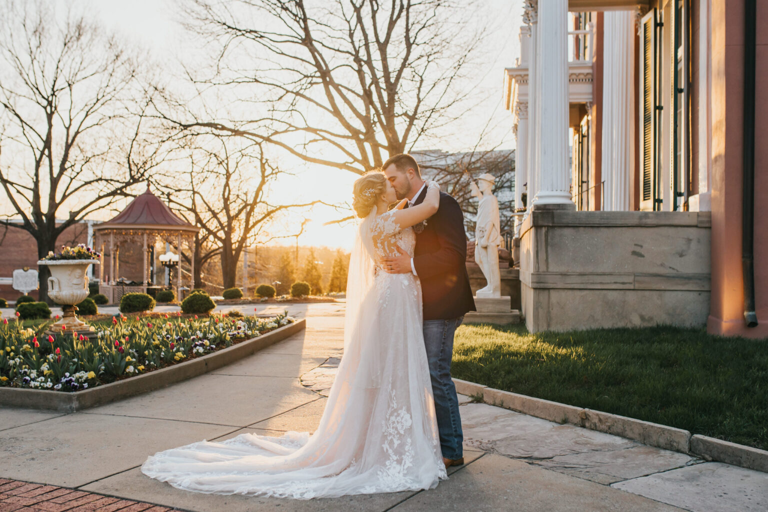 Elopement Wedding A newlywed couple, fresh from their elopement, embraces outside at sunset in front of an ornate building. The bride wears a white gown with a long train, and the groom sports a dark suit with jeans. Surrounding them are flowerbeds with blooming flowers, trees without leaves, and a gazebo in the background. Elopements Inc