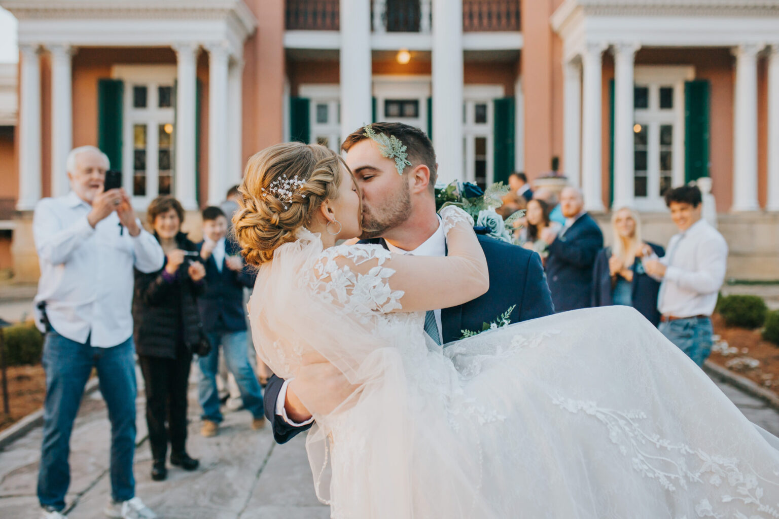 Elopement Wedding A groom in a navy suit carries his bride in a lace wedding dress and veil as they kiss in front of a peach-colored, columned building. Guests in the background, dressed in various attire, applaud and take photos, capturing the joyous moment of their elopement. Elopements Inc