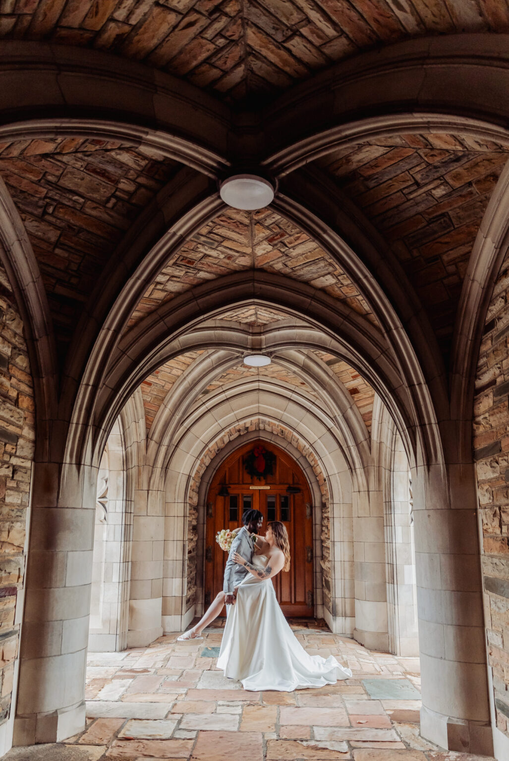 Elopement Wedding A newlywed couple kisses under an elegant stone archway, the elopement adding a touch of spontaneity to their special day. The bride, in a flowing white gown, holds a bouquet of yellow flowers, while the groom dips her in a romantic embrace. The archway's intricate brickwork leads to a large wooden door adorned with a circular wreath. Elopements Inc