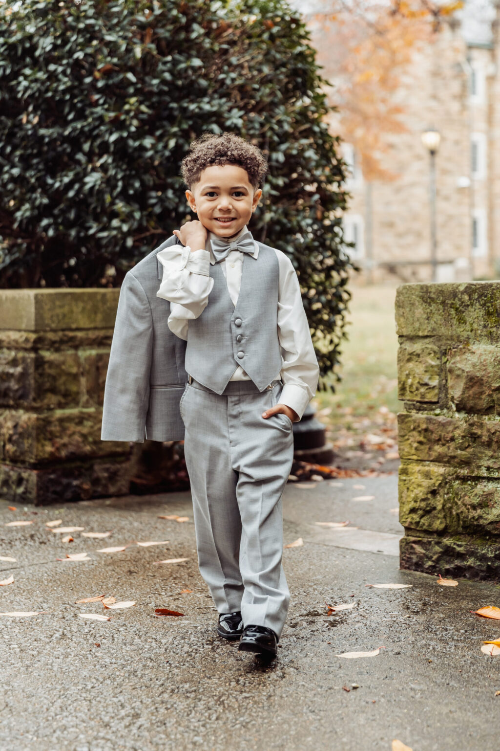 Elopement Wedding A young boy with curly hair walks outdoors, holding a gray suit jacket over his shoulder. He wears a matching gray vest, pants, and a bow tie over a white dress shirt. He smiles confidently, with one hand in his pocket. The background features stone walls, a bush, and autumn foliage perfect for an elopement. Elopements Inc