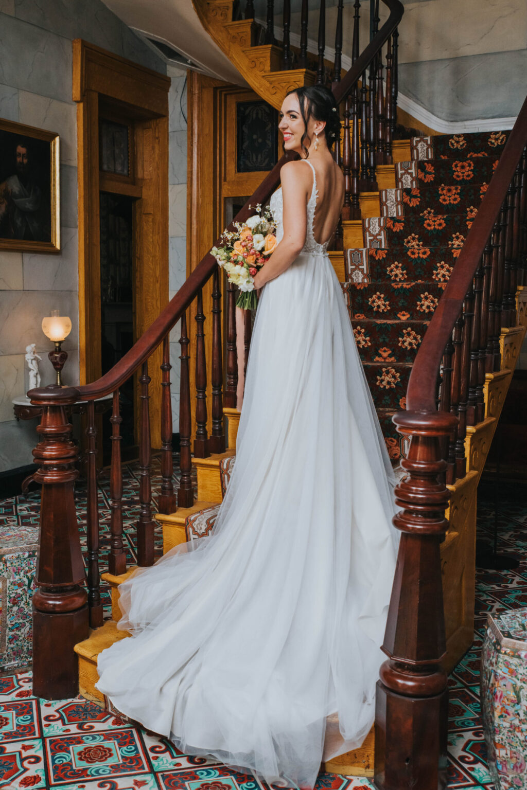 Elopement Wedding A bride, in a flowing white gown with a low back, stands on a grand staircase holding a bouquet of flowers. She looks over her shoulder, smiling. The staircase has ornate wooden railings and patterned carpet. The setting appears vintage with framed portraits and elaborate tiled flooring. Elopements Inc