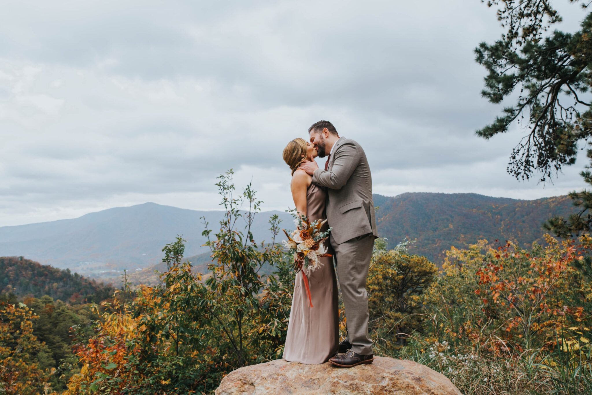 Elopement Wedding A couple embraces while standing on a large rock, surrounded by lush greenery and overlooking a mountain range. The woman, wearing a light pink dress and holding a bouquet of flowers, kisses the man, who is dressed in a light gray suit. The sky is cloudy, and the foliage is tinged with autumn colors. Elopements Inc