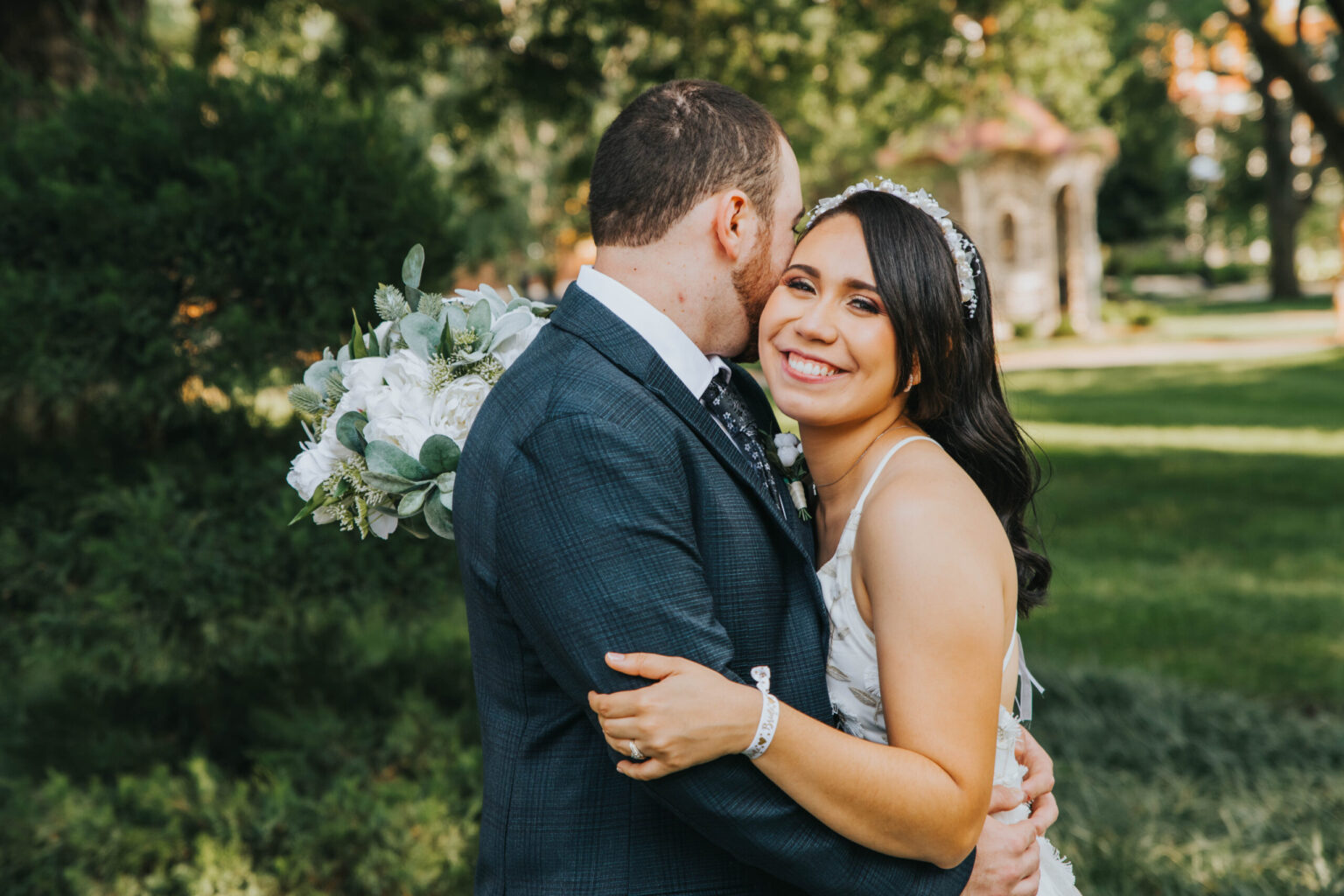 Elopement Wedding A bride and groom are embracing outdoors in front of a lush, green backdrop with trees and a gazebo. The bride, smiling and holding a bouquet of white flowers, wears a white dress and tiara. The groom, in a dark suit, has his back to the camera, his head close to the bride’s. Elopements Inc