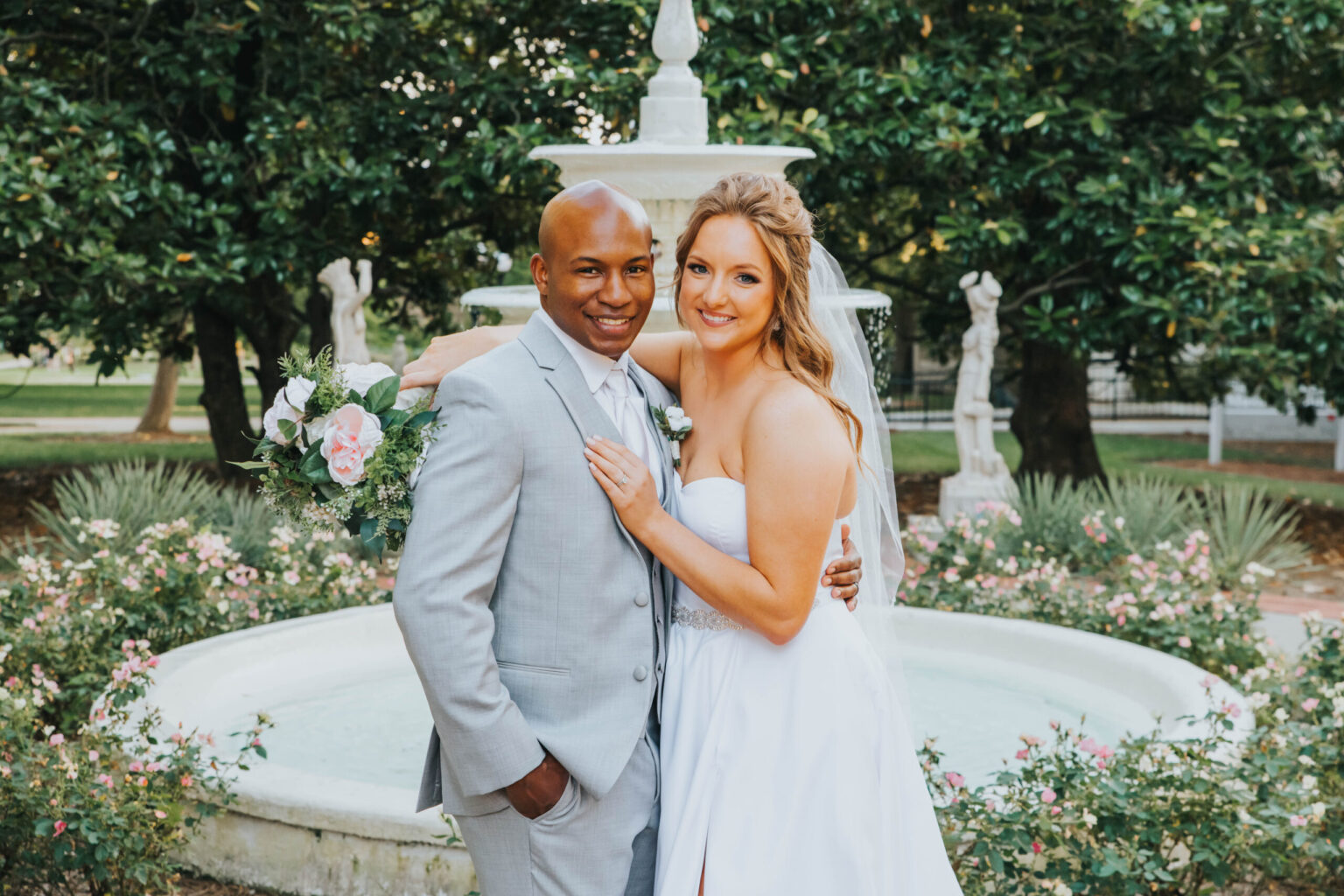 Elopement Wedding A newlywed couple poses in front of a fountain amidst lush greenery. The groom, dressed in a light gray suit, smiles while the bride, in a strapless white gown, holds a bouquet of pink and white flowers. They stand close, radiating happiness on their wedding day. Elopements Inc