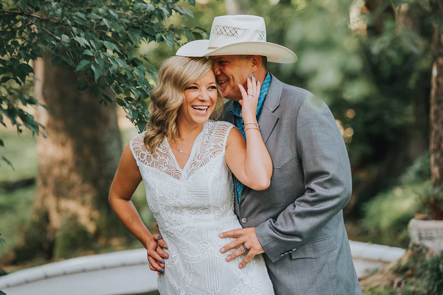 Elopement Wedding A joyful couple stands outdoors, embraced. The woman, in a lace white dress, has wavy blonde hair and laughs heartily with one hand on her hip. The man, wearing a light gray suit, light blue shirt, and a cowboy hat, smiles warmly and holds her around the waist. Trees and greenery surround them. Elopements Inc