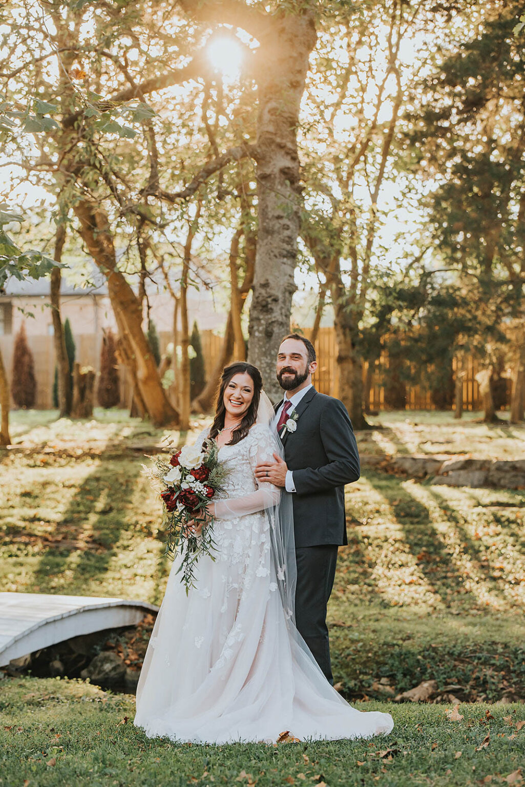 Elopement Wedding A bride and groom stand together outdoors under a tree with sunlight filtering through its branches. The bride wears a white lace gown and holds a bouquet of red and white flowers. The groom is in a dark suit with a white shirt and maroon tie. They both smile, with greenery and a wooden fence in the background. Elopements Inc