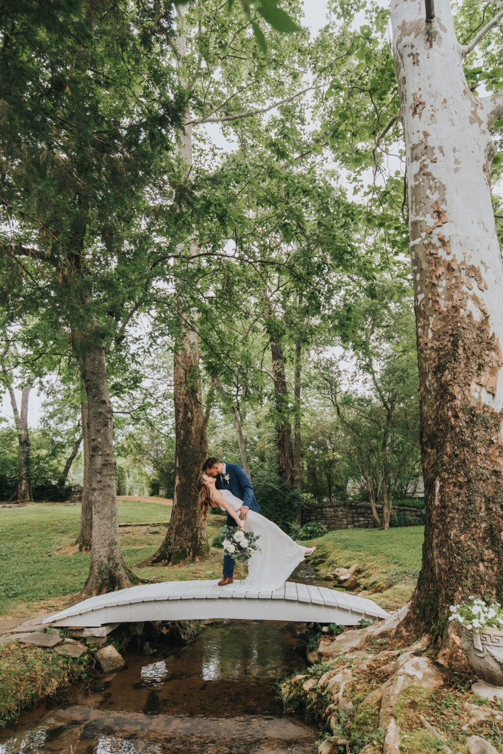 Elopement Wedding A bride and groom share a kiss on a small arched wooden bridge over a creek in a lush green park. The bride holds a bouquet of flowers and is dressed in a white gown, while the groom wears a blue suit. Tall trees with green foliage surround them, adding a natural, serene backdrop to the scene. Elopements Inc