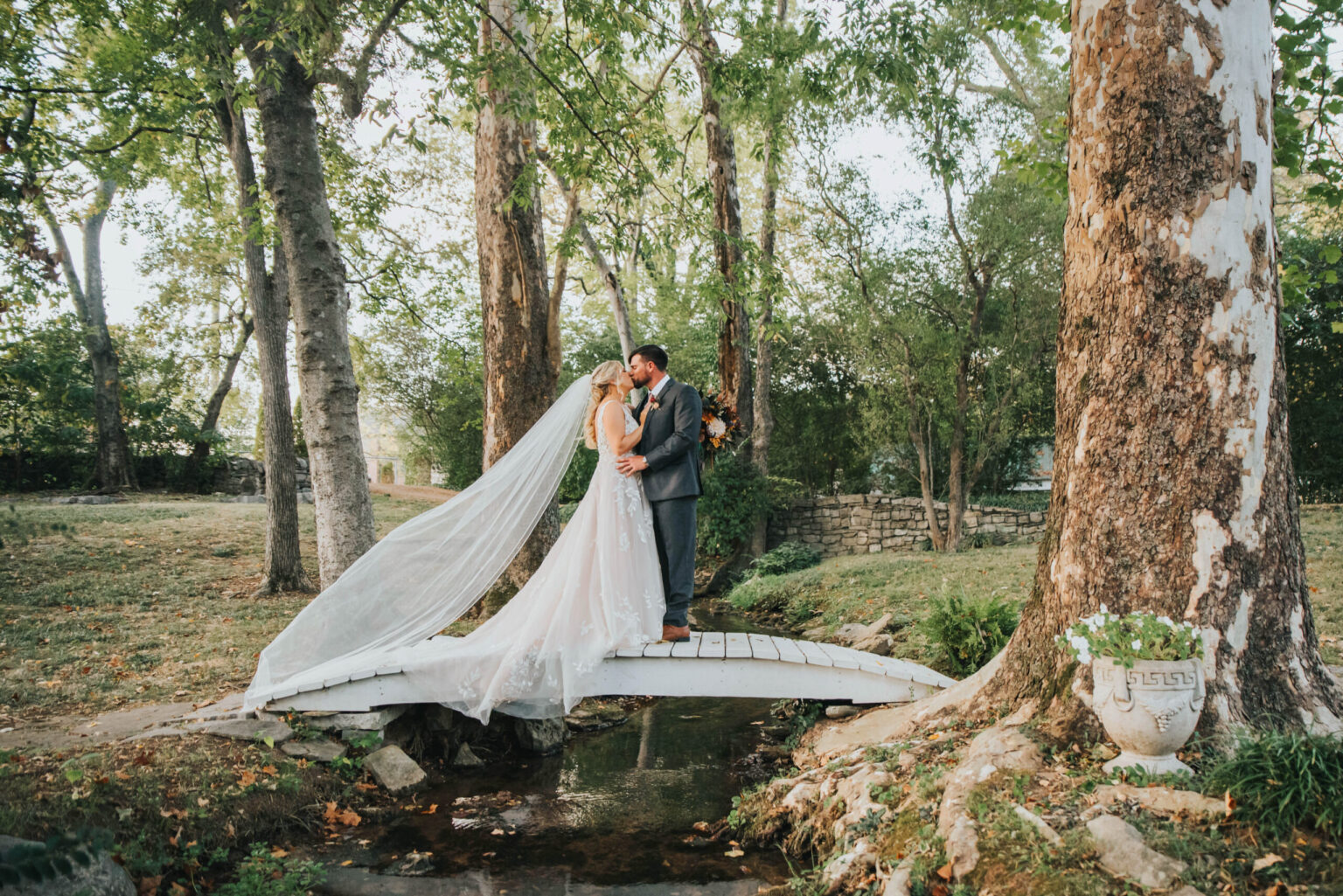 Elopement Wedding A bride and groom kiss on a small wooden bridge surrounded by lush greenery and large trees. The bride's flowing veil drapes over the bridge, and they are both elegantly dressed in wedding attire. A decorative planter with flowers is visible to the right, near the base of a tree. Elopements Inc