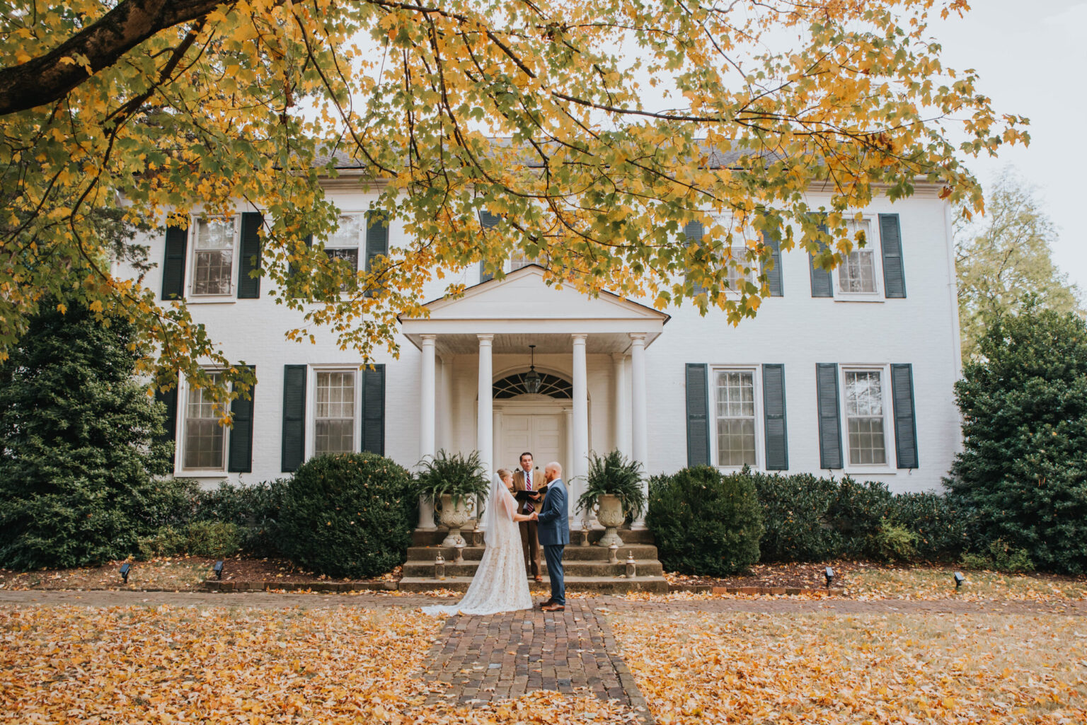 Elopement Wedding A bride and groom stand under a tree with yellow leaves in front of a large, white, colonial-style house with green shutters. The bride is wearing a white dress and holding a bouquet, and the groom is in a dark suit. They are embracing each other, surrounded by autumn foliage. Elopements Inc