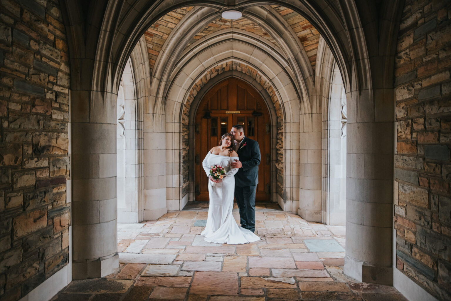 Elopement Wedding A newlywed couple stands under an arched stone hallway. The bride, in a white gown, holds a bouquet of flowers while the groom, in a dark suit, embraces her. They stand in front of a wooden double door with gothic-style architecture, illuminated by soft, natural light from outside. Elopements Inc