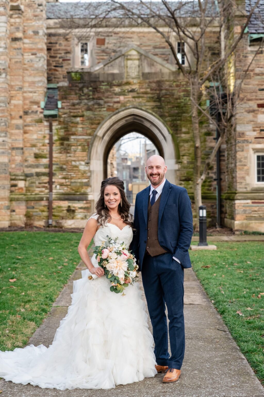 Elopement Wedding A bride in a white gown holds a bouquet of flowers, standing beside a groom in a navy blue suit with a brown vest. They pose in front of a stone archway with ivy and a green lawn, smiling at the camera on their wedding day. The background features a stone building with tall windows. Elopements Inc