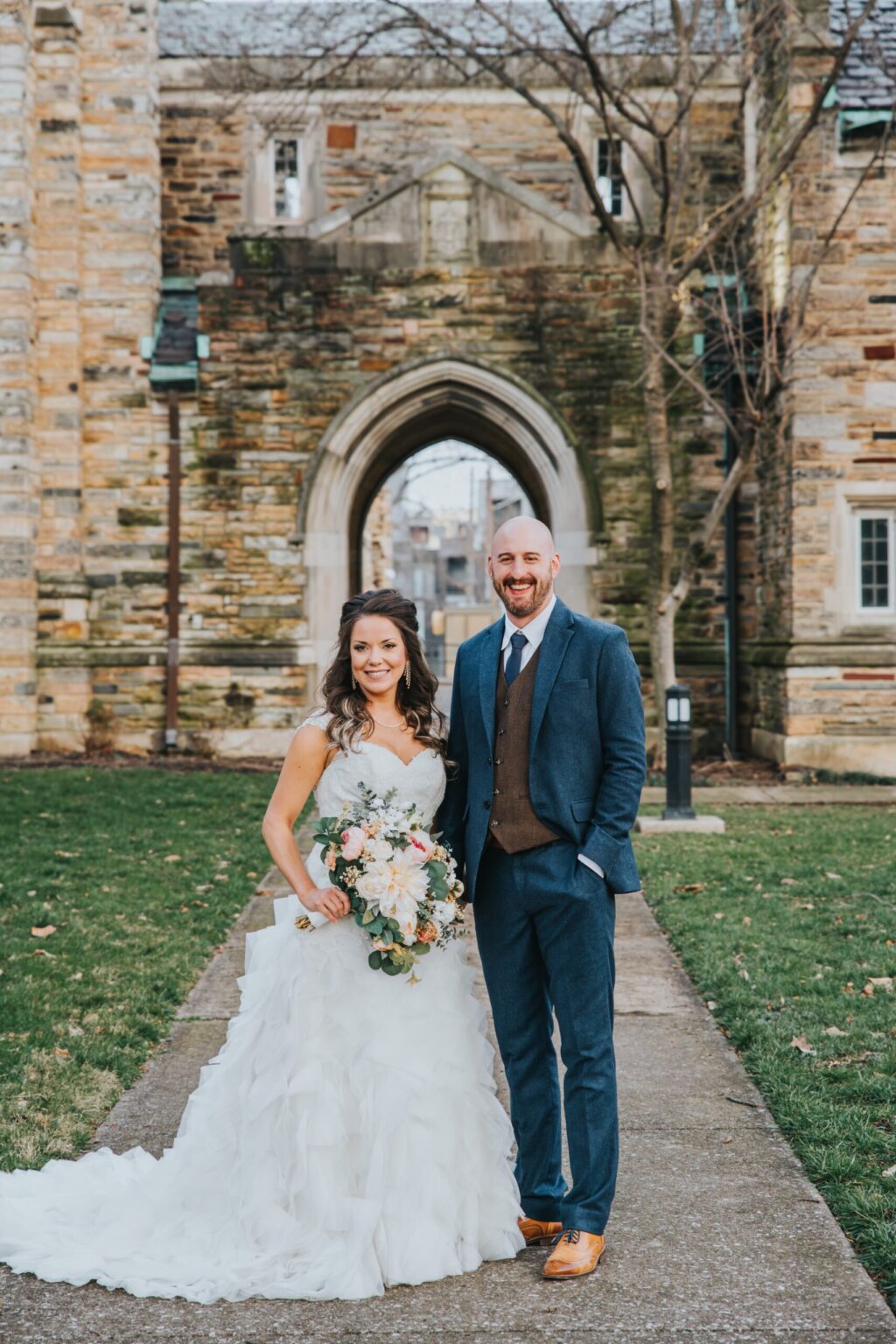 Elopement Wedding A bride and groom stand smiling in front of a stone archway and building. The bride is in a white, ruffled gown and holding a bouquet of flowers. The groom is in a blue suit with a brown vest and tie. They are standing on a pathway with grass on both sides. Leafless trees are visible in the background. Elopements Inc