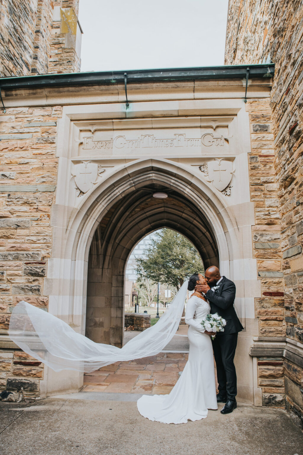 Elopement Wedding A bride and groom share an intimate moment under a stone archway. The bride is in a long white dress with a flowing veil, holding a bouquet of white flowers. The groom is in a black suit. They stand close, gazing into each other's eyes as the veil billows gracefully in the breeze. Elopements Inc