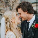 Elopement Wedding A joyful bride and groom stand close, about to kiss. The bride, in a lace wedding gown with a long, white veil, has blonde, wavy hair. The groom wears a black suit with a white shirt, black tie, and a vibrant red rose boutonnière. They are outside in front of a stone building. Elopements Inc