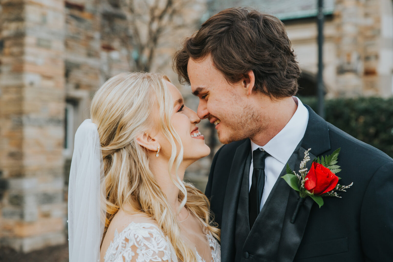 Elopement Wedding A joyful bride and groom stand close, about to kiss. The bride, in a lace wedding gown with a long, white veil, has blonde, wavy hair. The groom wears a black suit with a white shirt, black tie, and a vibrant red rose boutonnière. They are outside in front of a stone building. Elopements Inc