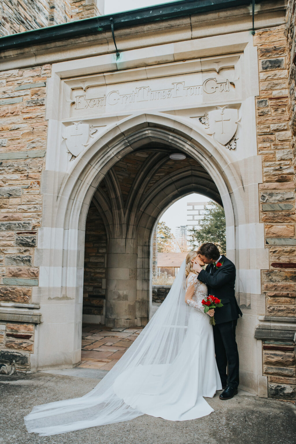 Elopement Wedding A newlywed couple embraces under a stone archway. The bride, in a long white dress and veil, holds a bouquet of red roses. The groom, wearing a black suit and red boutonniere, kisses her forehead. The archway is part of a building with tan and brown stone, featuring an inscription above. Elopements Inc