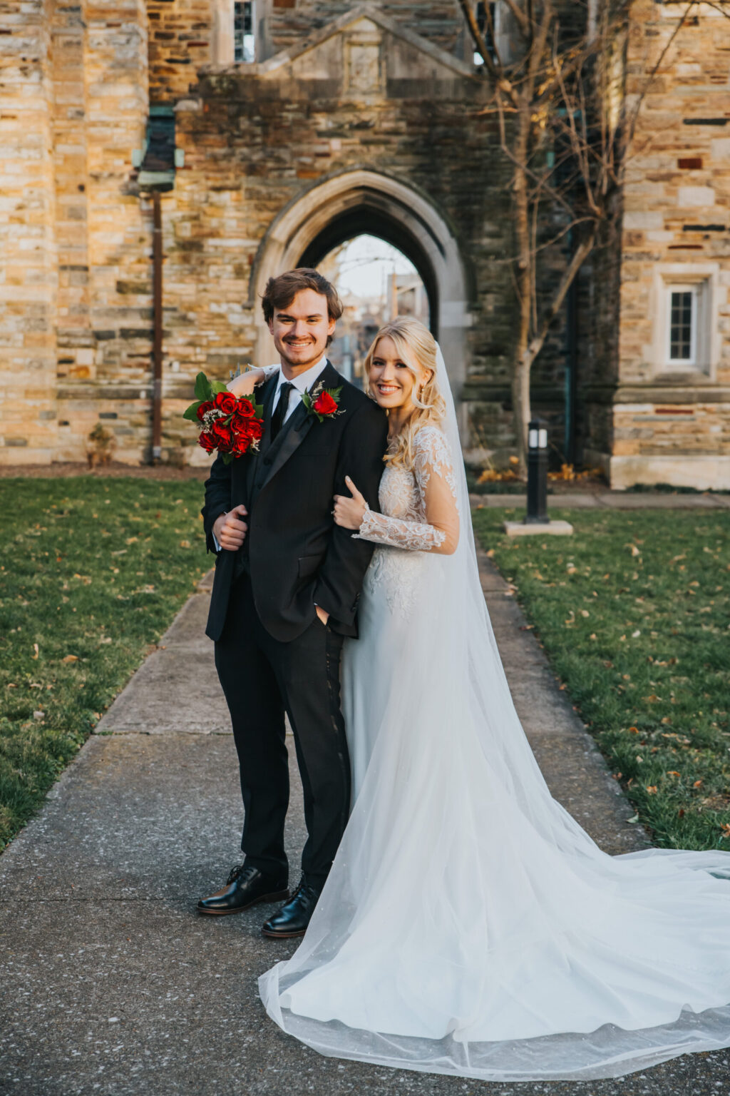 Elopement Wedding A bride and groom stand outdoors in front of a stone building with an archway. The bride, in a long white dress with lace sleeves, smiles as she holds the groom's arm. The groom, in a black suit and tie, smiles while holding a bouquet of red flowers. The ground is paved with a small grass area nearby. Elopements Inc