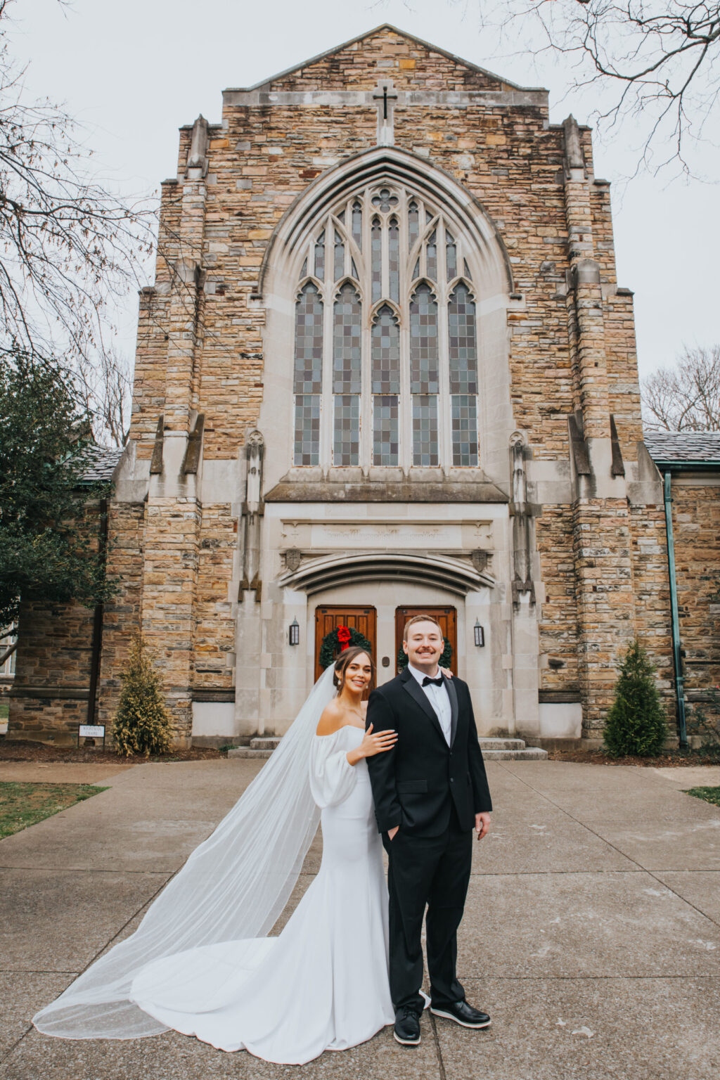 Elopement Wedding A bride and groom stand in front of a large stone church with arched windows and a wooden door. The bride wears a white dress and veil, and the groom is dressed in a black suit with a bow tie. Both are smiling and holding hands. The church has gothic architectural elements. Elopements Inc