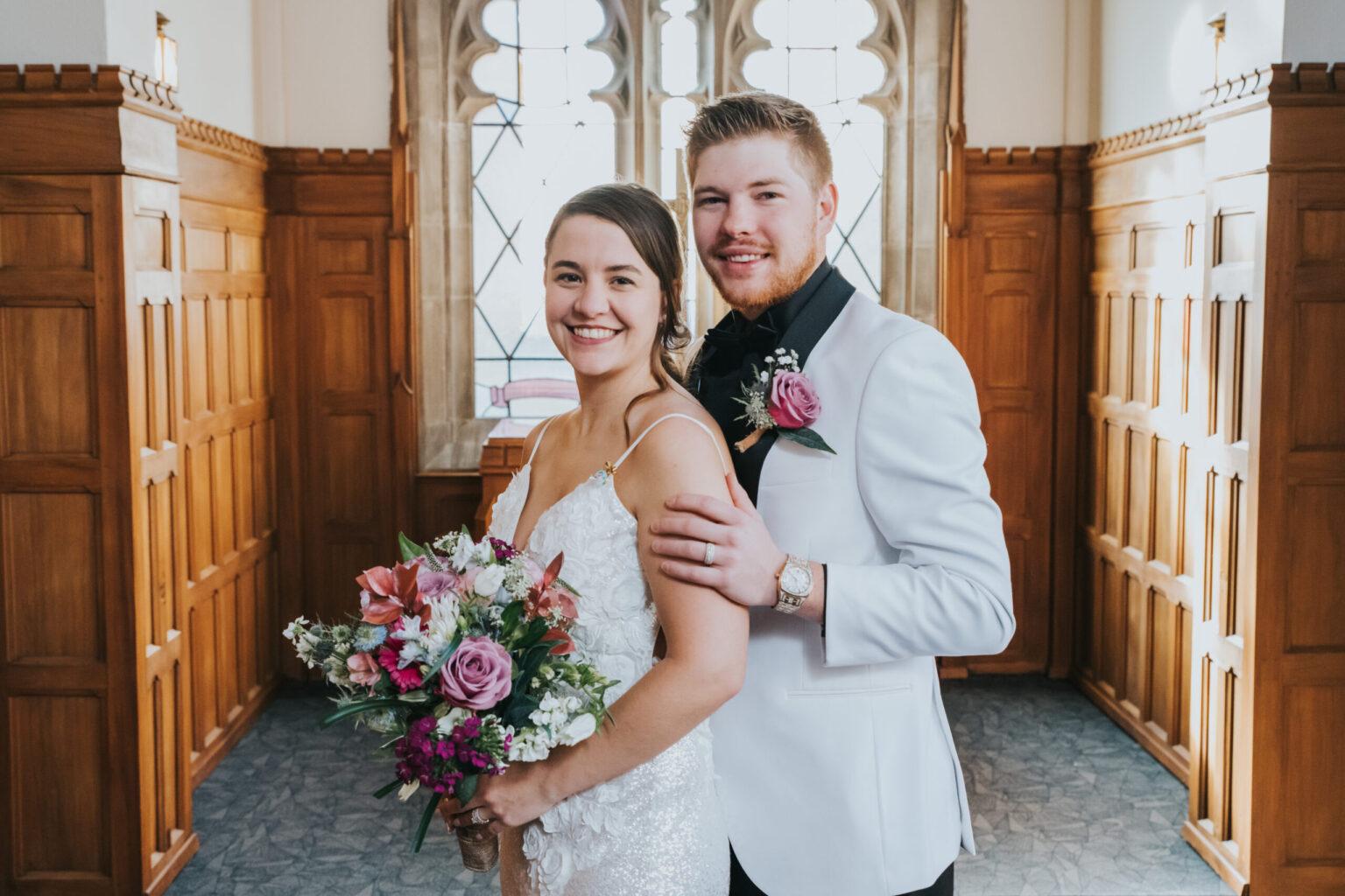 Elopement Wedding A couple poses for a wedding photo in a wood-paneled room with arched windows. The bride wears a white lace dress and holds a colorful bouquet. The groom, in a white tuxedo jacket with a black bow tie and a pink boutonniere, stands behind her, resting his hand on her shoulder. Both are smiling. Elopements Inc