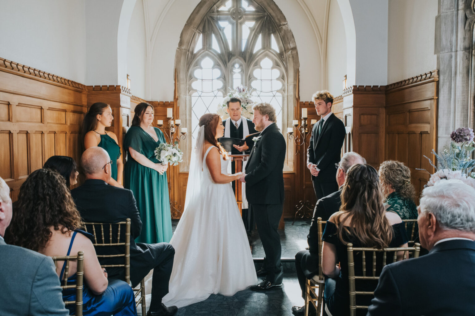 Elopement Wedding A bride and groom stand facing each other, holding hands, in a small chapel with wooden paneling and a large arched window. Bridesmaids in green dresses and groomsmen in black suits stand beside them. Guests are seated on golden chairs, facing the couple. An officiant is speaking to them. Elopements Inc