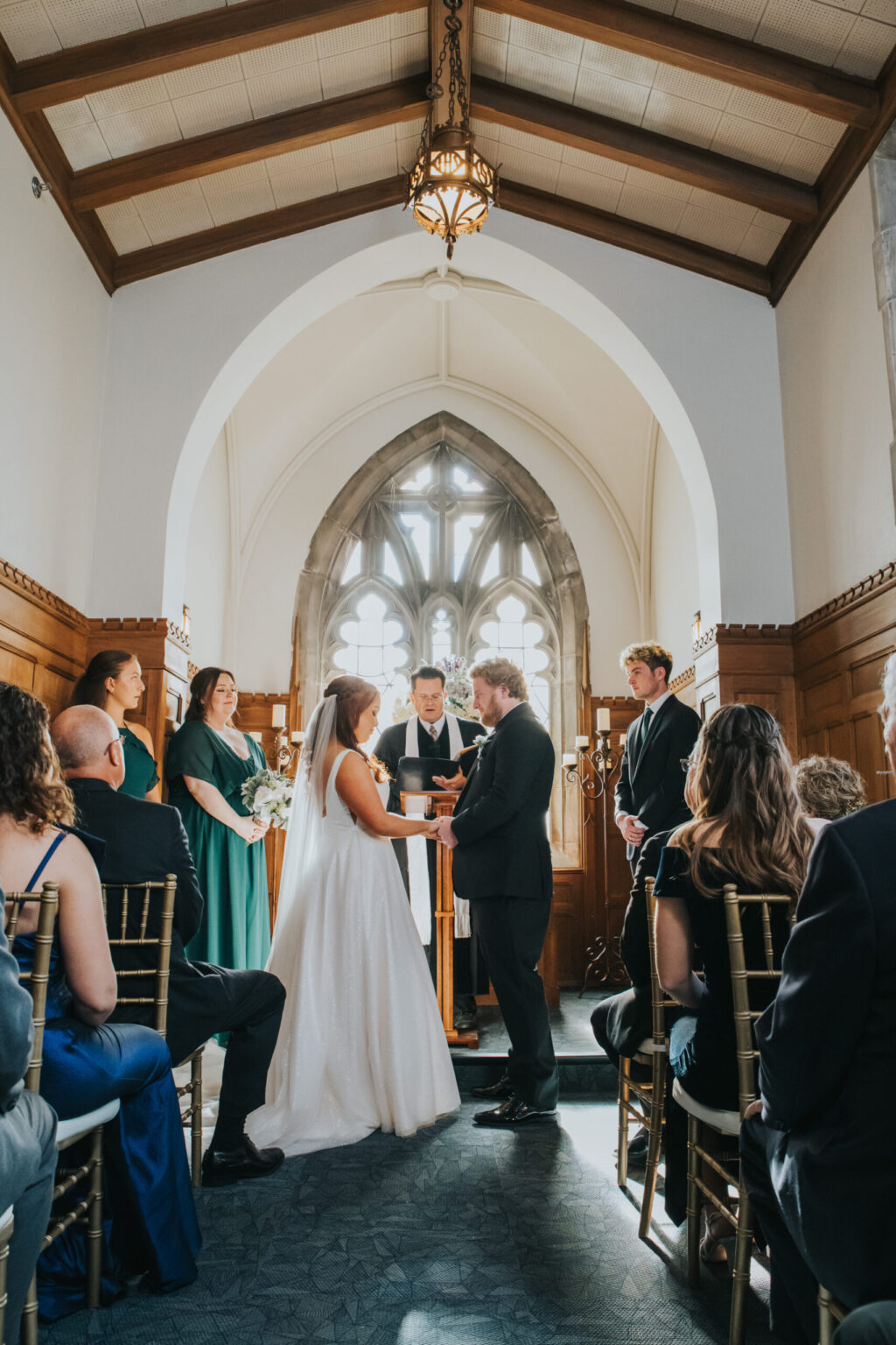 Elopement Wedding A bride and groom stand hand-in-hand at the altar during a wedding ceremony in a small chapel with a high arched ceiling and gothic windows. The officiant reads from a book. Bridesmaids in green dresses and groomsmen in black suits stand on either side, and guests are seated facing the couple. Elopements Inc