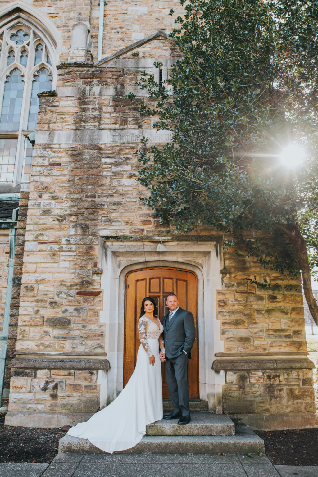 Elopement Wedding A bride in a long-sleeve white gown and a groom in a gray suit stand hand in hand in front of an arched wooden door set in a brick church wall. The setting sun shines through the tree on the right, casting a warm light over the couple and the aged brick facade. Elopements Inc