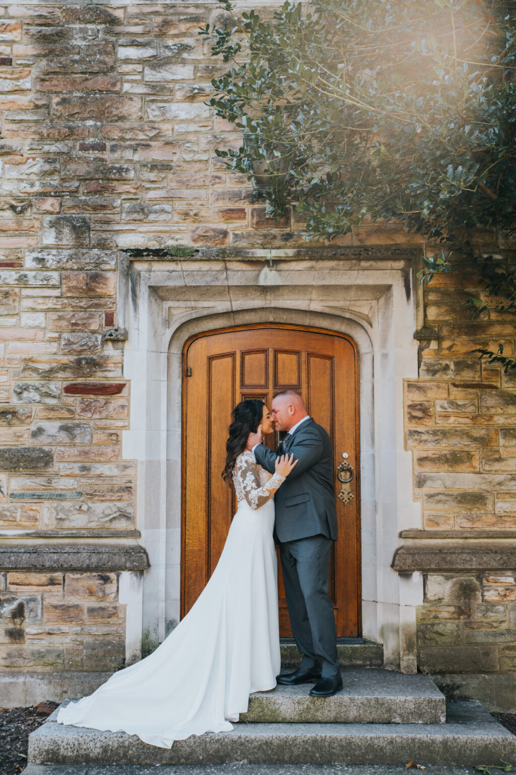 Elopement Wedding A bride and groom share a kiss in front of an arched wooden door set into a stone wall. The bride wears a long white gown with lace sleeves and has long dark hair. The groom is dressed in a gray suit. They stand on stone steps, framed by greenery on the right and sunlight illuminating the scene. Elopements Inc