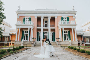 Elopement Wedding A bride and groom share a kiss in front of a large, elegant, peach-colored mansion with tall white columns, green shutters, and statues decorating the facade. The bride, in a white gown, holds a bouquet as the groom, in a black suit, dips her slightly backwards. Manicured gardens surround them. Elopements Inc