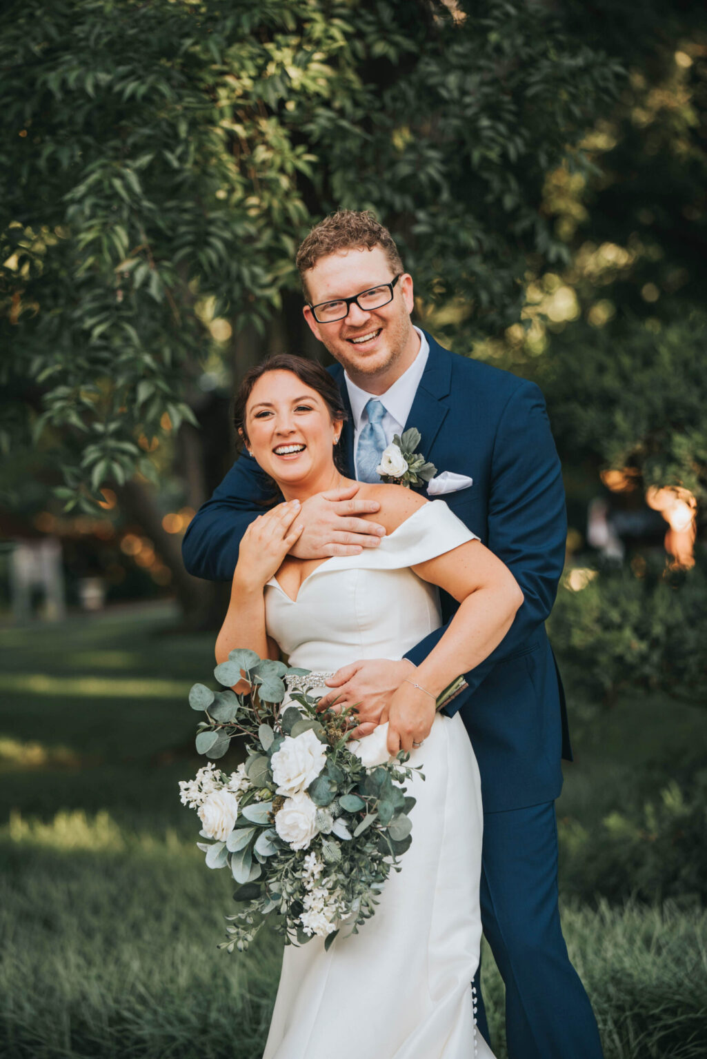 Elopement Wedding A joyful pair is shown in their wedding attire. The bride, wearing an off-shoulder white gown and holding a bouquet of white flowers and greenery, is embraced from behind by the groom, who is in a dark blue suit, light blue tie, and glasses. They stand in a lush, green outdoor setting. Elopements Inc