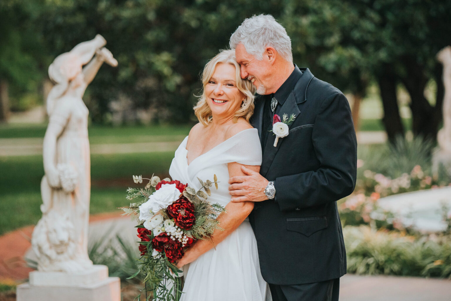 Elopement Wedding A mature couple stands together outdoors at what appears to be a wedding. The woman, wearing a white dress and holding a bouquet of red and white flowers, smiles brightly. The man, dressed in a black suit, stands close beside her with his arm affectionately around her shoulder. Statues are visible in the background. Elopements Inc