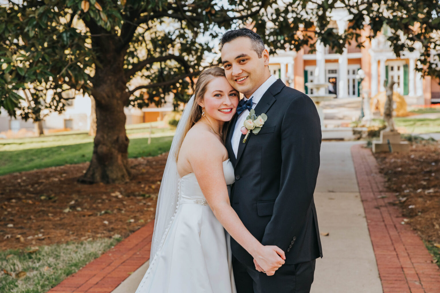 Elopement Wedding A bride in a white dress and veil stands beside a groom in a black suit and tie. They are holding hands and smiling while standing on a brick pathway in an outdoor park-like setting with trees and a large, historic building in the background. Elopements Inc