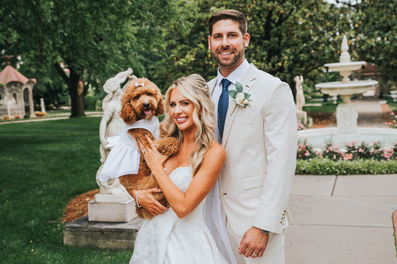 Elopement Wedding A bride and groom are standing outdoors in a park with lush greenery. The bride, in a white strapless wedding gown, holds a small brown dog dressed in a white outfit. The groom, in a light gray suit with a blue tie and white boutonnière, stands beside her. A fountain and statues are in the background. Elopements Inc