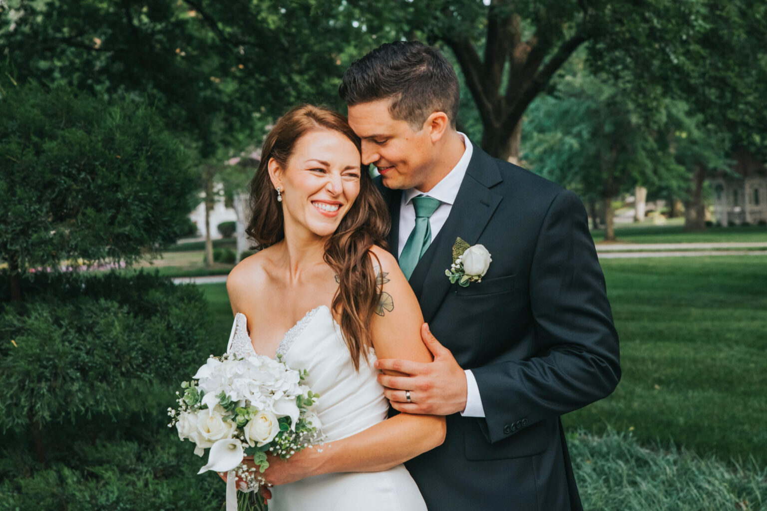 Elopement Wedding A bride in a white dress and a groom in a black suit with a green tie stand outdoors in a leafy garden. The bride holds a bouquet of white flowers and laughs while the groom, smiling, stands behind her with his arms wrapped around her waist. Both look joyful and content. Elopements Inc