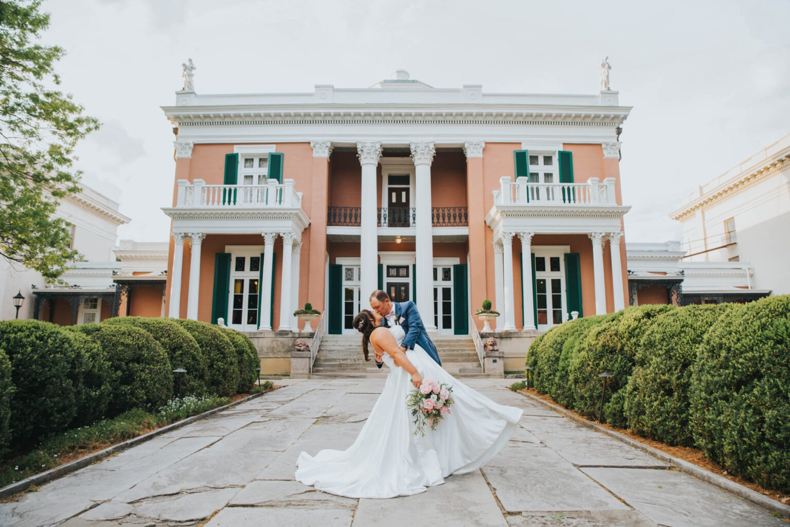 Elopement Wedding A couple dressed in wedding attire share a kiss while standing on a pathway bordered by greenery. The groom, in a blue suit, dips the bride, who wears a flowing white gown and holds a bouquet. They are in front of a large, elegant mansion with columns and balconies. Elopements Inc