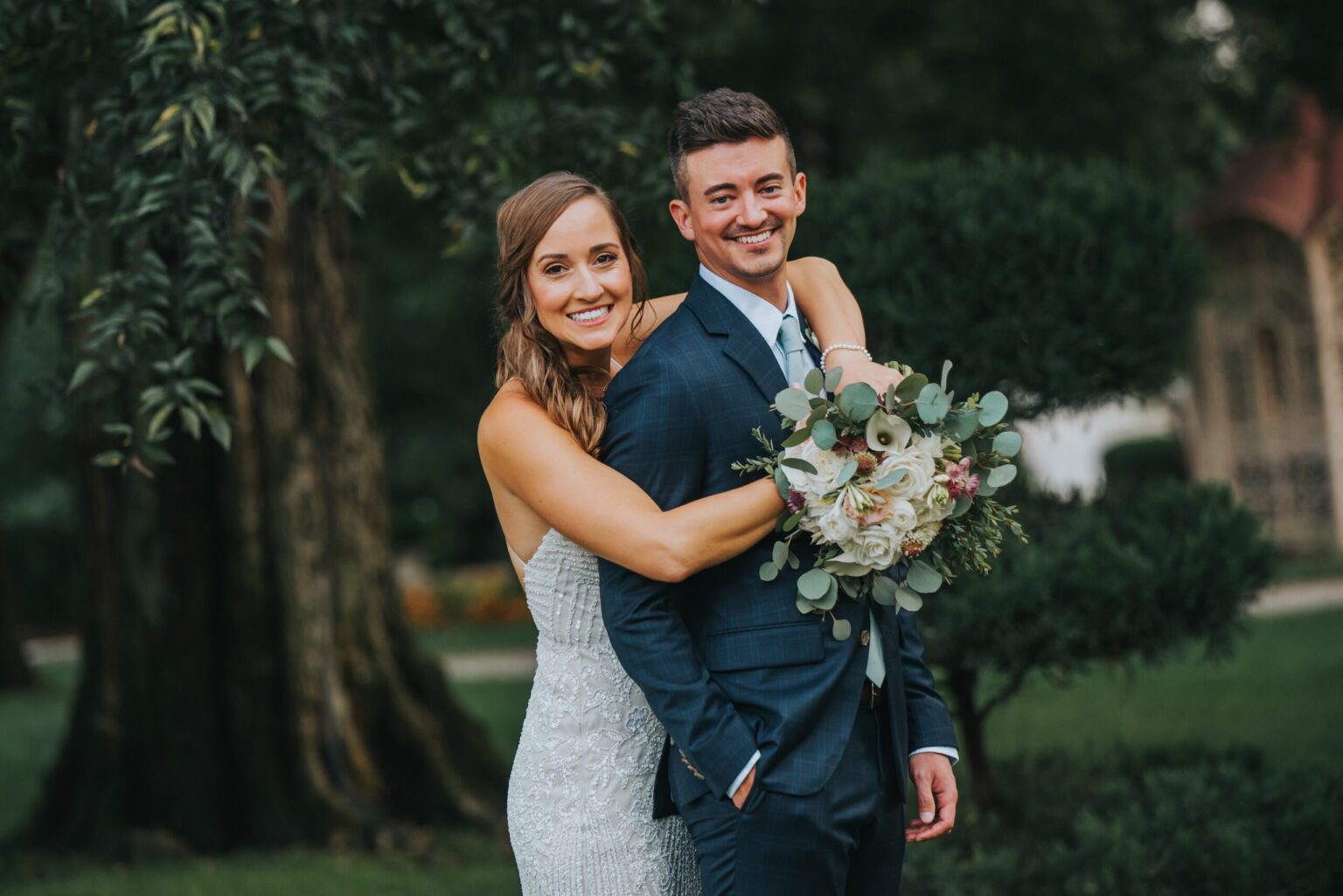 Elopement Wedding A cheerful bride and groom pose outdoors in front of a tree. The bride, in a white strapless dress, hugs the groom from behind while smiling. The groom, in a blue suit and tie, smiles warmly. The bride holds a bouquet of white and pink flowers with green foliage. They are surrounded by greenery. Elopements Inc