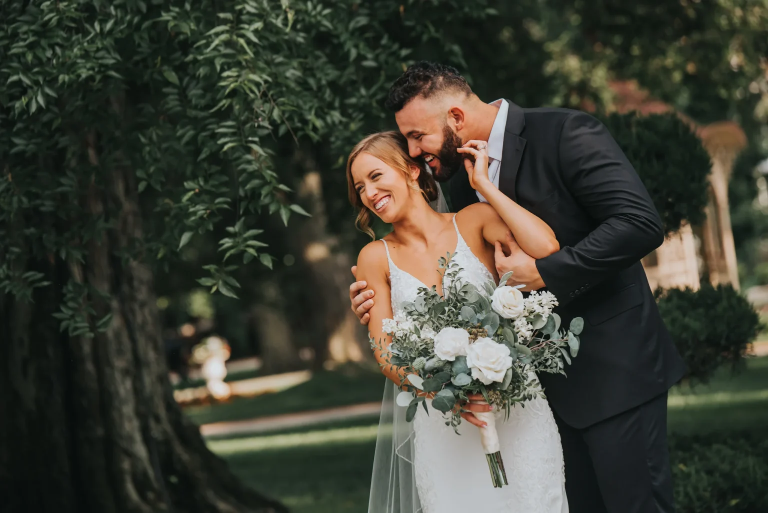 Elopement Wedding A joyful bride in a white lace gown holds a bouquet of white and green flowers while smiling and leaning into her groom, who is in a black suit. They are standing outdoors next to a large tree on a sunny day with greenery in the background. Elopements Inc