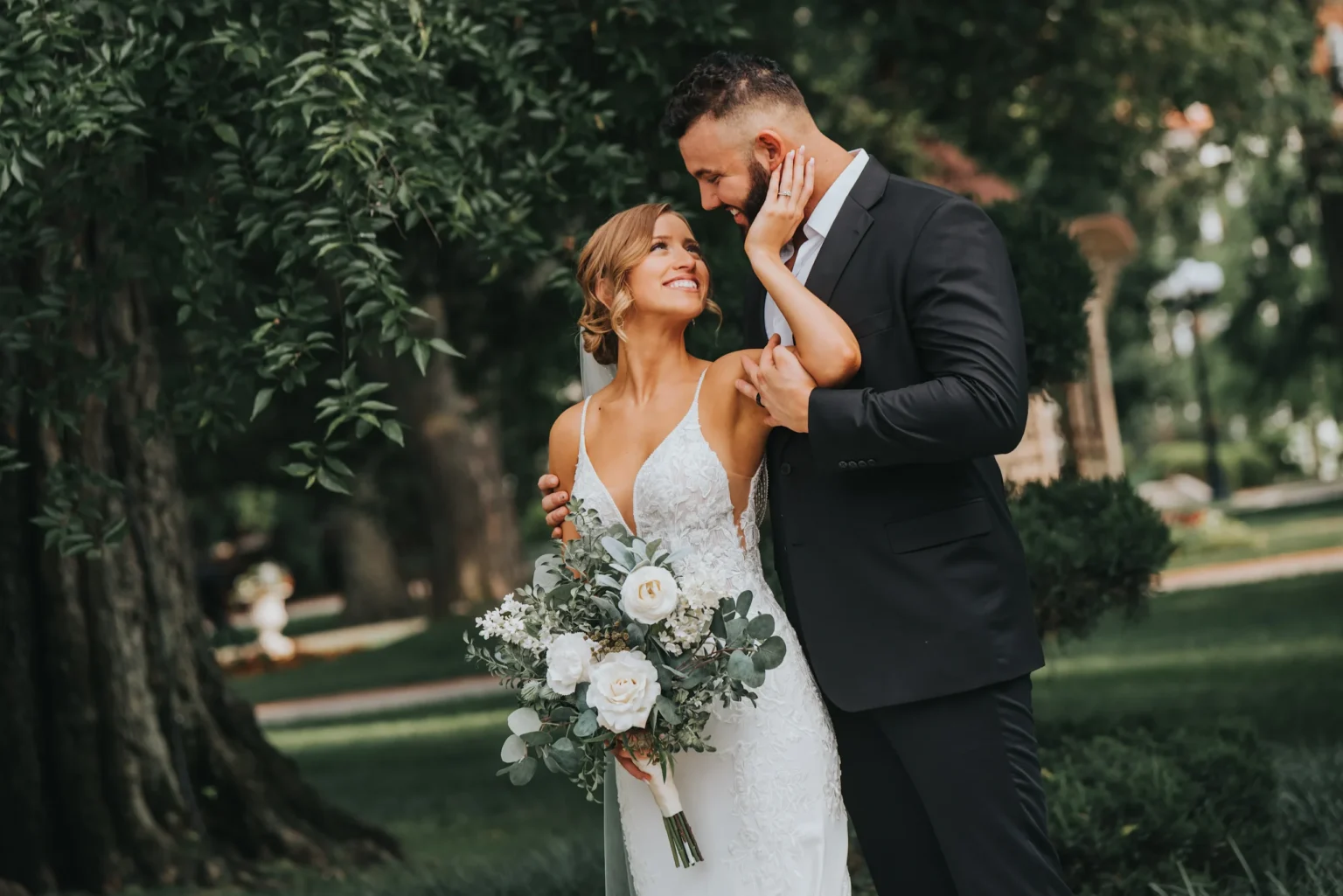 Elopement Wedding A bride in a white lace dress holding a bouquet of white roses and greenery gazes up lovingly at the groom in a black suit and tie. They stand closely in a lush, green outdoor setting with trees and a manicured garden in the background. The groom gently touches the bride's face. Elopements Inc