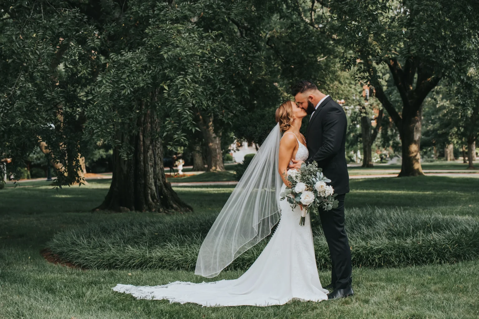 Elopement Wedding A bride and groom stand close under large trees in a lush green park. The bride, in a white dress with a long train and veil, holds a bouquet of white and blush flowers. The groom wears a black suit. Both appear to be touching their foreheads together, sharing an intimate moment. Elopements Inc