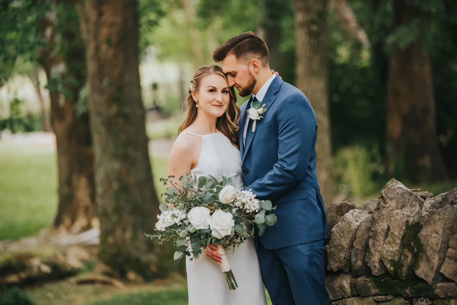 Elopement Wedding A bride in a sleeveless white wedding dress holds a bouquet of white flowers and greenery. She stands beside a groom in a blue suit and white shirt, who gently rests his head against hers. They are outdoors near a stone wall with green trees and grass in the background, creating a serene and romantic atmosphere. Elopements Inc