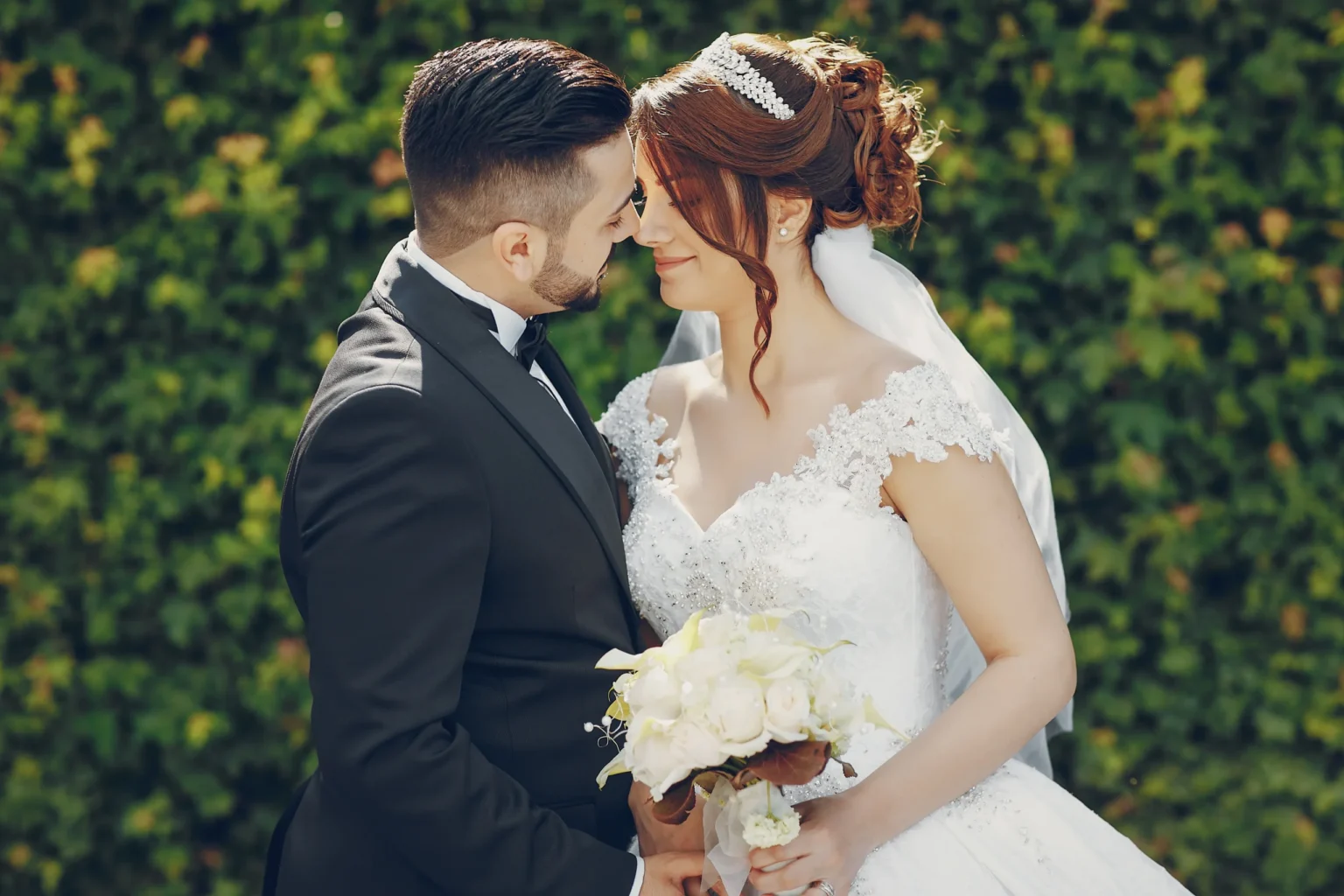 Elopement Wedding A bride and groom smile with closed eyes, touching foreheads. The groom wears a black tuxedo, and the bride dons a white lace wedding gown and tiara, holding a bouquet of white flowers. They stand in front of a leafy green background. Elopements Inc