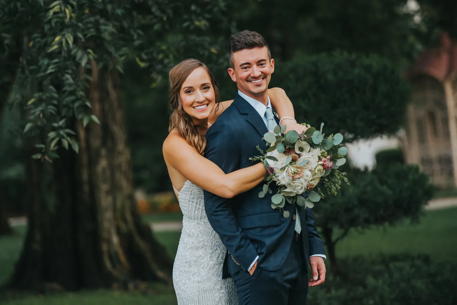 Elopement Wedding A smiling bride embraces a smiling groom from behind as they pose outdoors. The bride wears a strapless white gown and holds a bouquet of white and pastel flowers. The groom is dressed in a blue suit with a tie. They stand in a garden with lush greenery and trees visible in the background. Elopements Inc