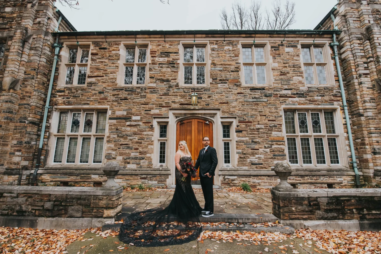 Elopement Wedding A couple stands holding hands in front of a stone building with arched wooden doors and large windows. The woman wears a long black dress with a flowing train, and the man is dressed in a dark suit. Fallen leaves are scattered on the ground around them, adding an autumnal feel to the scene. Elopements Inc