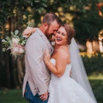 Elopement Wedding A laughing bride in a white wedding dress and veil embraces a groom in a beige suit jacket and jeans. She holds a bouquet of pink and white flowers. The couple stands outside, with a backdrop of green trees and soft sunlight filtering through the leaves. Elopements Inc