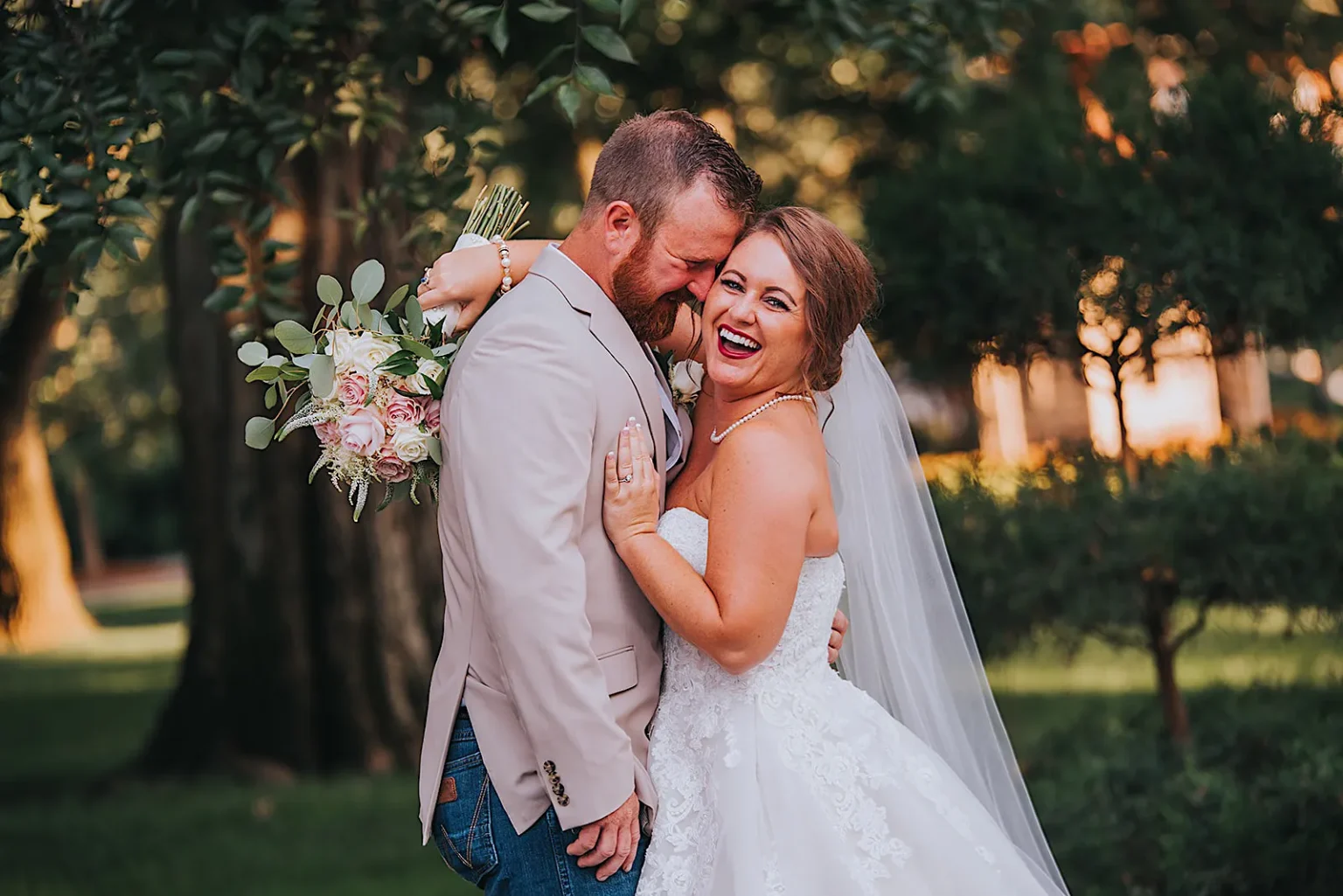 Elopement Wedding A laughing bride in a white wedding dress and veil embraces a groom in a beige suit jacket and jeans. She holds a bouquet of pink and white flowers. The couple stands outside, with a backdrop of green trees and soft sunlight filtering through the leaves. Elopements Inc