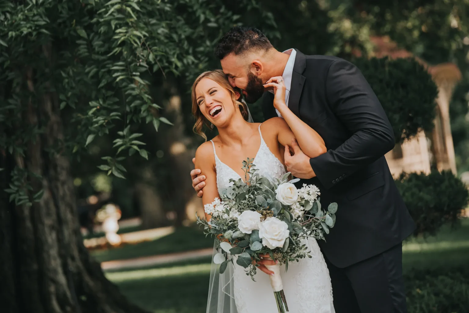 Elopement Wedding A joyful bride in a white lace wedding dress holds a bouquet of white roses and greenery, laughing as the groom, in a black suit, affectionately kisses her on the cheek. They stand in a lush, green outdoor setting with large trees in the background, creating a romantic ambiance. Elopements Inc