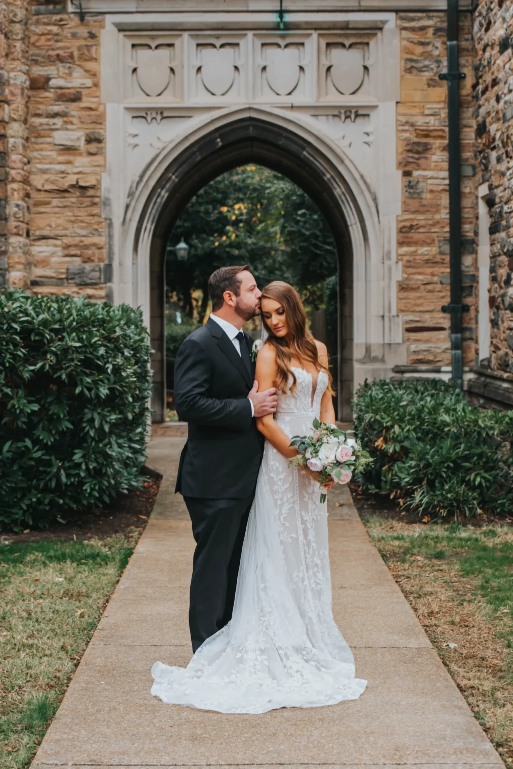 Elopement Wedding A groom in a black suit kisses a bride’s forehead as they stand outside in front of a gothic-style archway with stone walls. The bride is wearing a white lace gown and holding a bouquet of flowers. They are framed by greenery and a pathway leading towards the archway. Elopements Inc